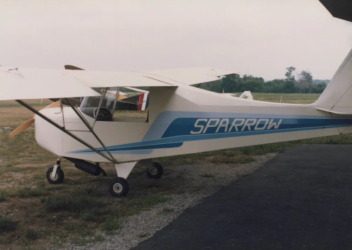 A white and blue Carlson Sparrow parked beside a taxiway near a hangar