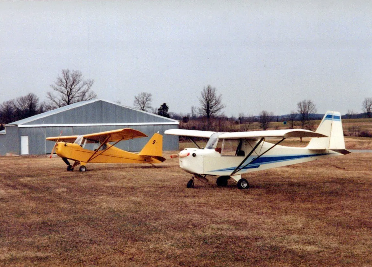A yellow and a white Carlson Sparrow parked side by side on a grass airfield