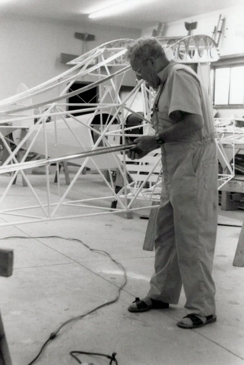 Black-and-white photograph of a man working on welded steel-tube fuselage frames in a shop