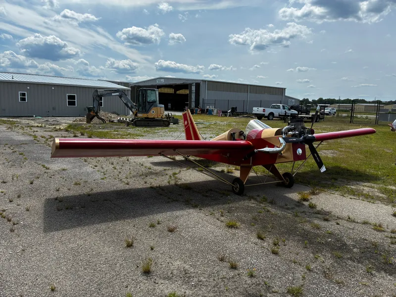 A red Mini-Max aircraft with a bare wood-and-fabric fuselage parked on a cracked asphalt ramp in front of hangars