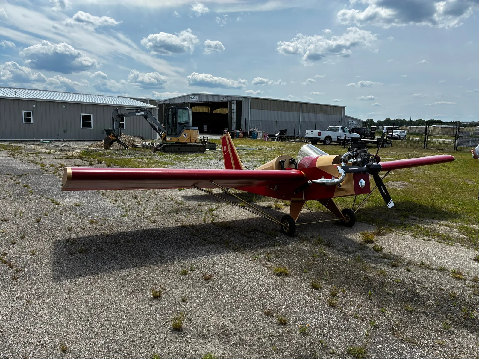 A red Mini-Max aircraft with a bare wood-and-fabric fuselage parked on a cracked asphalt ramp in front of hangars