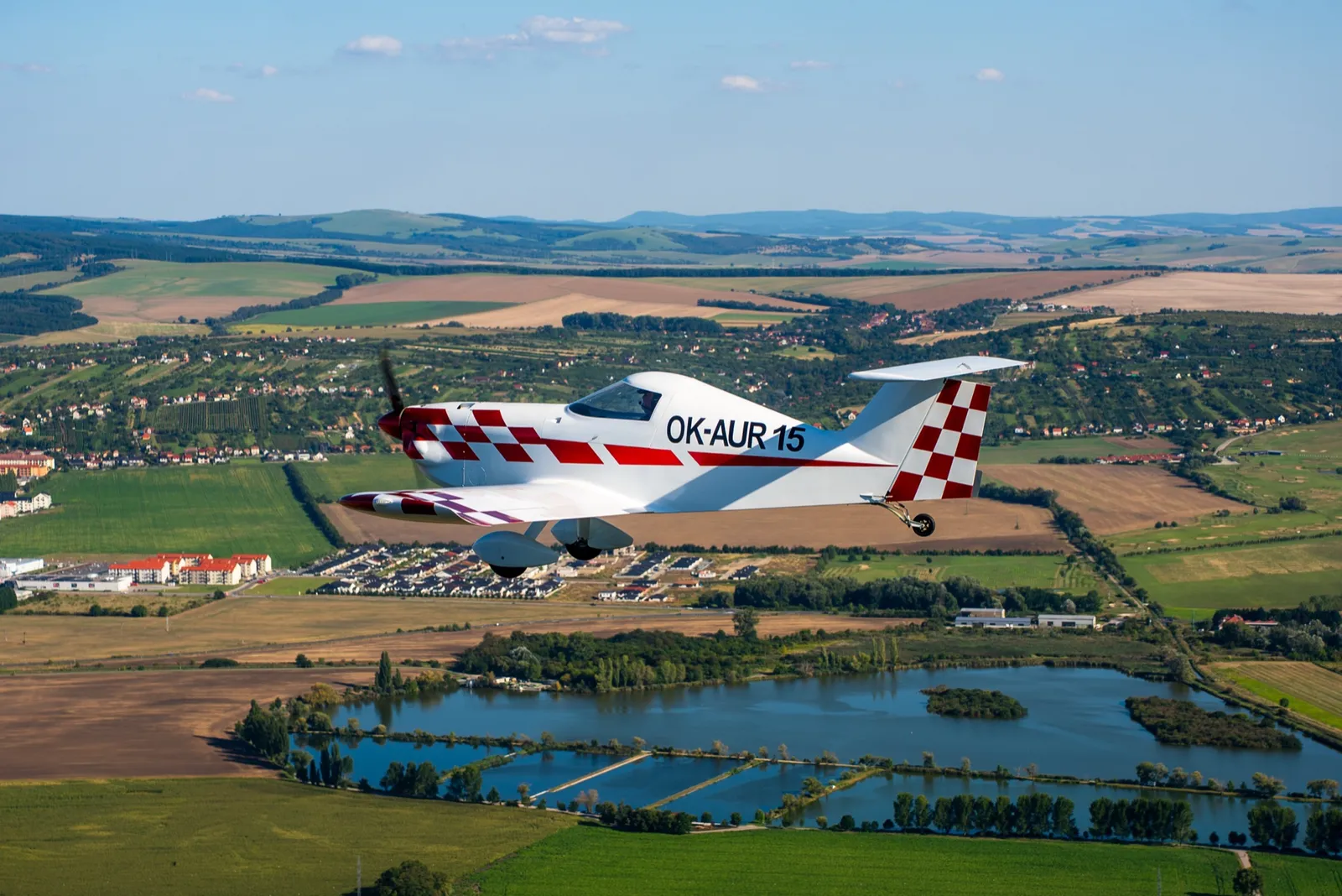 A white and red single-seat SD-1 Minisport banking over fields, lakes and a village