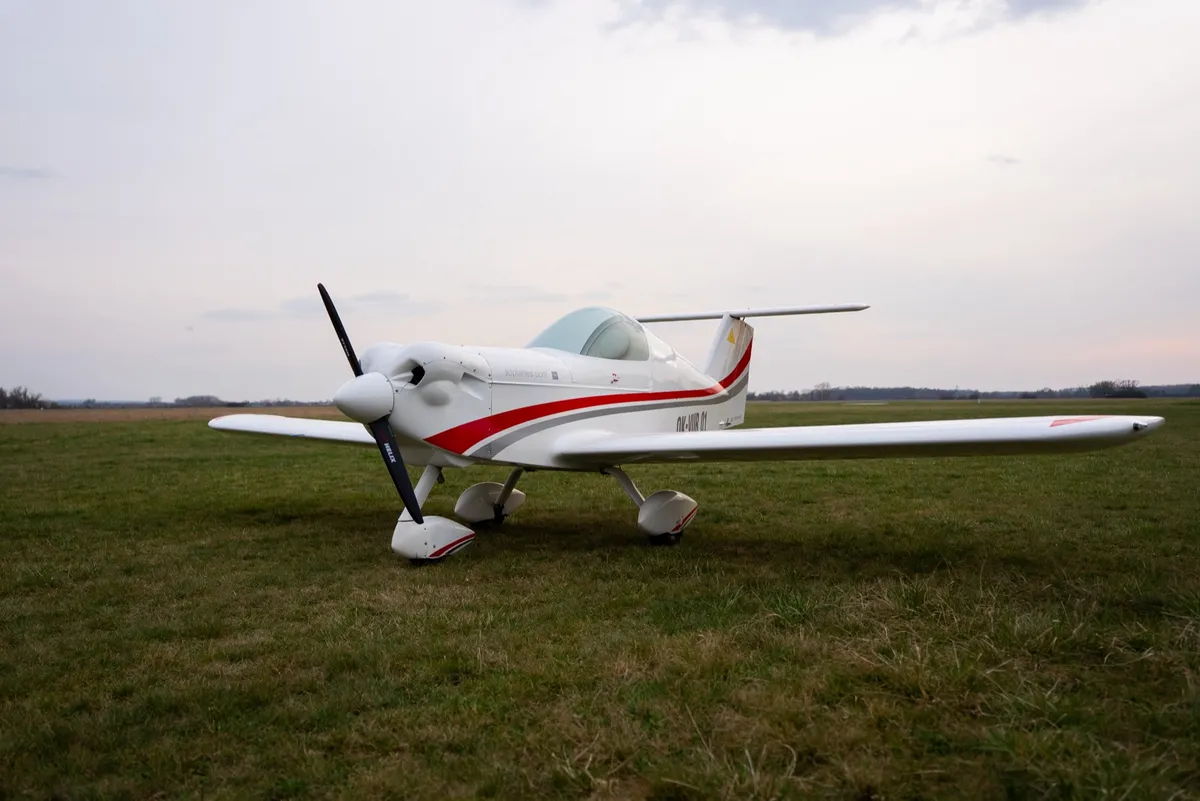 A white SD-1 Minisport with red trim parked on grass, seen from the front quarter with the propeller stopped